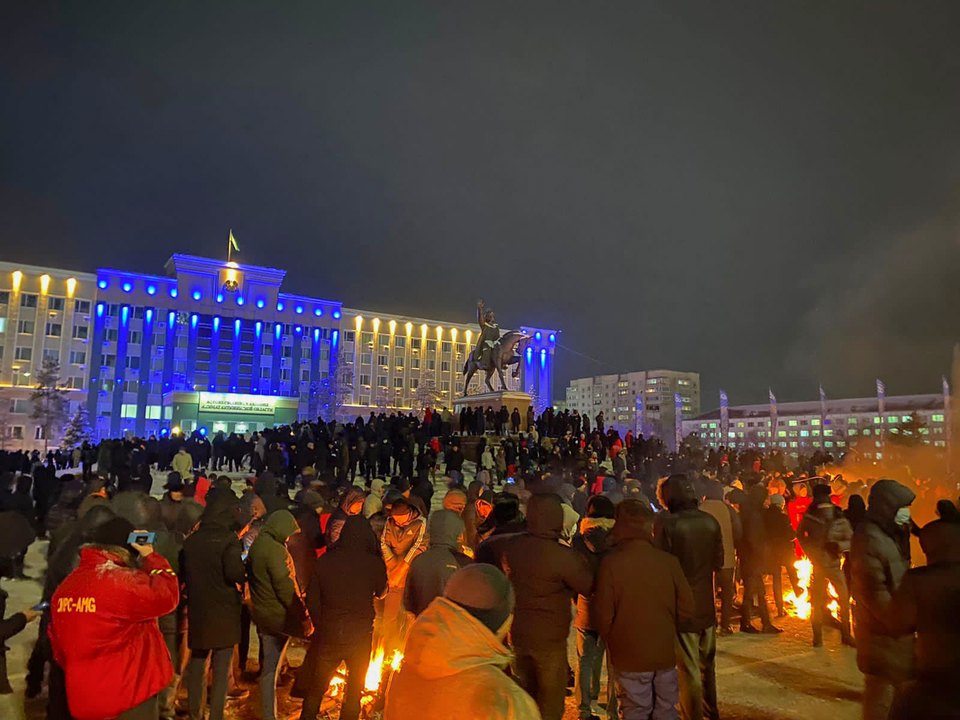 Люди на заснеженной площади перед освещённым зданием и памятником; на переднем плане горят костры.